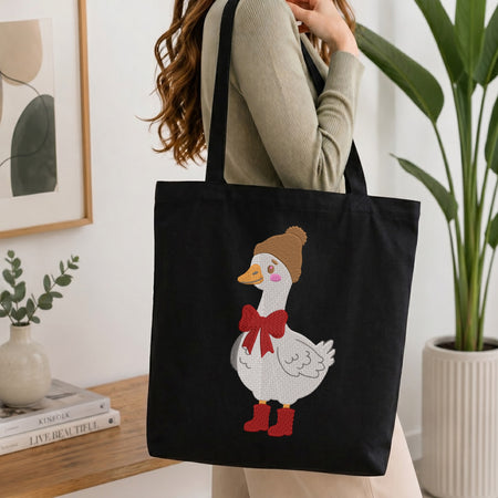A person wearing a beige ribbed sweater carries a black canvas tote bag with a white goose graphic wearing a brown beanie, red bow, and red boots, in a bright indoor setting with a plant and books in the background.