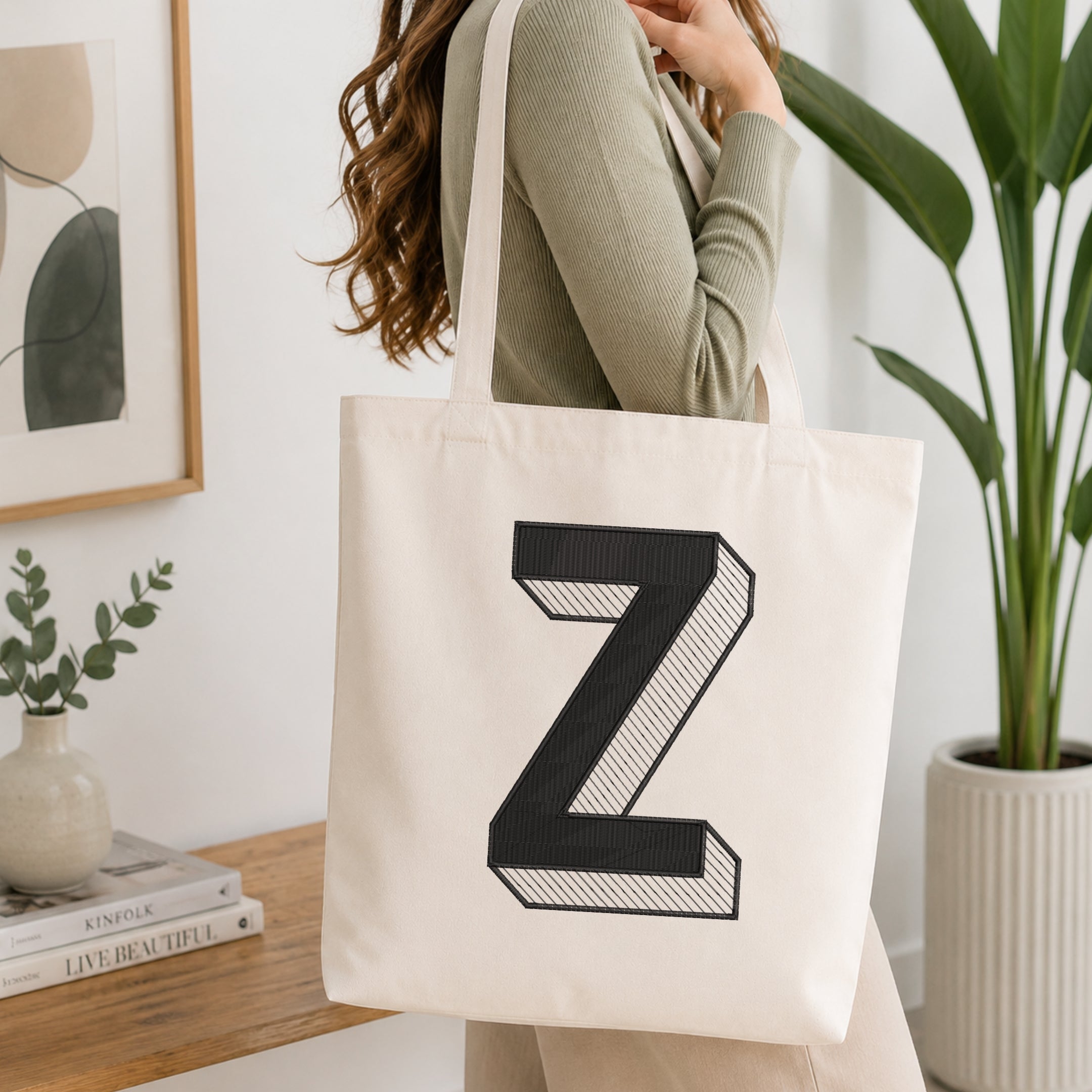 A woman wearing a sage green ribbed long-sleeve top carries a cream canvas tote bag with a large black 3D-effect letter Z printed on the front, set in a bright, modern room with a wooden console table, framed art, and a potted plant.