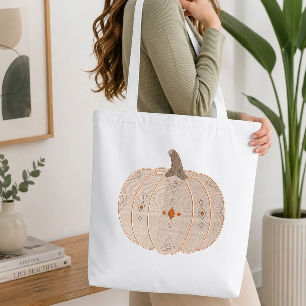 Woman carrying a white natural canvas tote bag featuring an embroidered geometric pumpkin design in a bright, modern interior setting.   