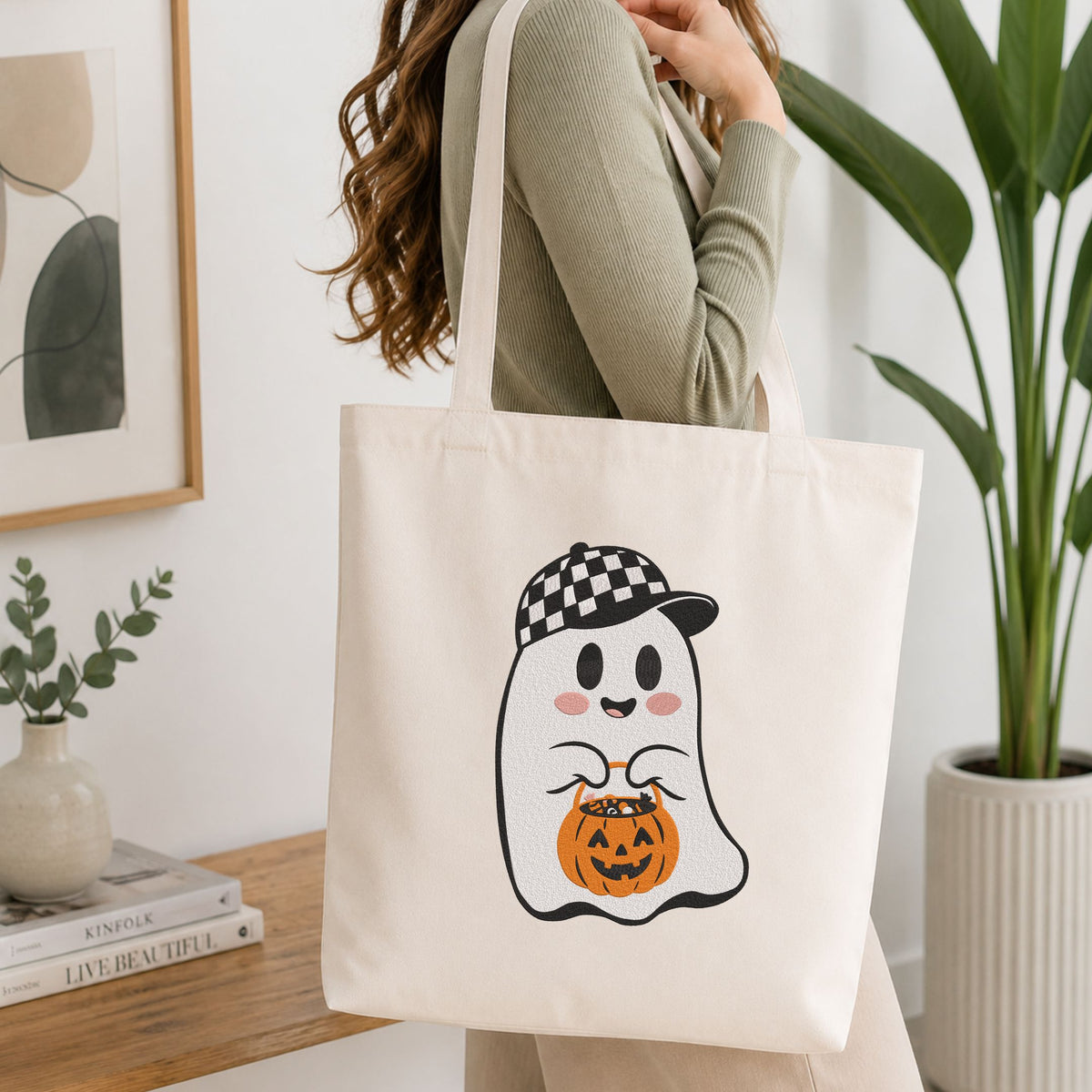  Woman carrying a white natural canvas tote bag featuring a cute ghost wearing a checkered baseball cap holding a pumpkin treat bucket machine embroidery design in a bright, modern interior setting   