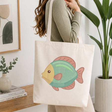 A woman wearing a sage green ribbed sweater carries a cream canvas tote bag with a colorful cartoon fish graphic. The background is a bright, modern room with a wooden table, books, a small plant, and a large green potted plant.