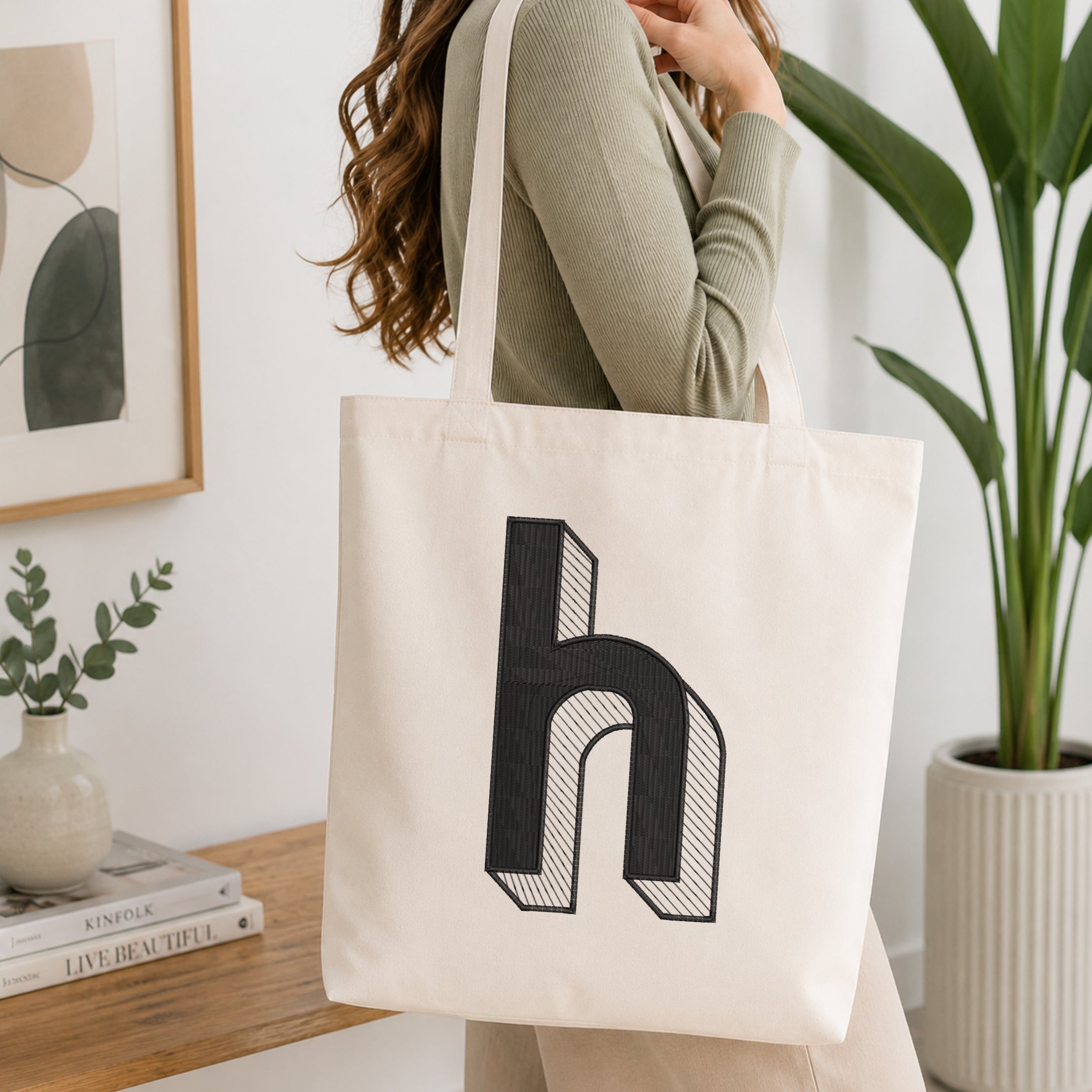 A woman wearing a sage green ribbed long-sleeve top carries a cream canvas tote bag with a large black stylized letter 'h' printed on the front, set in a bright, modern room with a wooden table, framed art, and a potted plant.