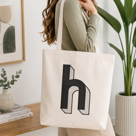 A woman wearing a sage green ribbed long-sleeve top carries a cream canvas tote bag with a large black stylized letter 'h' printed on the front, set in a bright, modern room with a wooden table, framed art, and a potted plant.