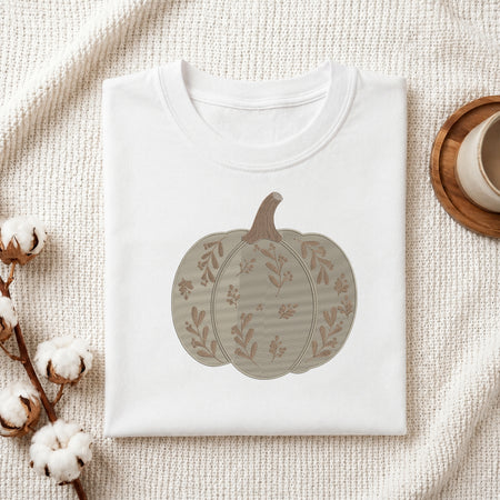 A folded white crewneck t-shirt with a printed autumn pumpkin design, styled on a textured cream fabric with cotton bolls and a wooden cup in the background.