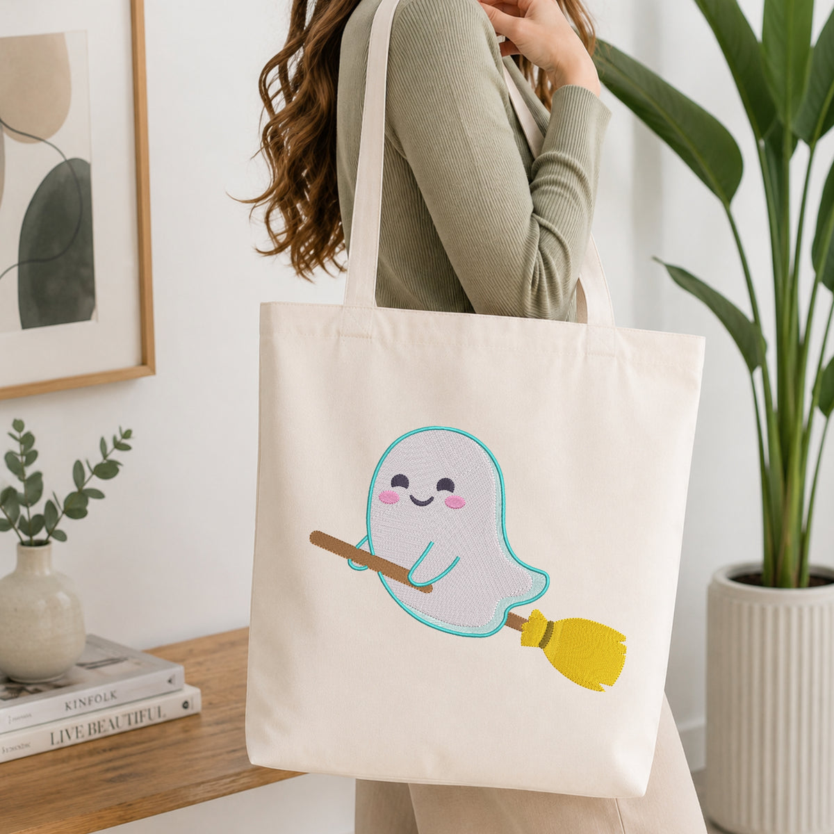 A woman holds a cream canvas tote bag with a cute embroidered ghost holding a yellow broomstick. The scene is set in a bright, modern room with a wooden table, books, and a potted plant in the background.