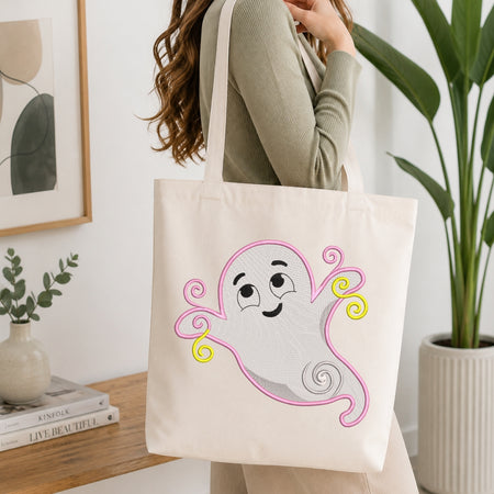 An off-white canvas tote bag with a cute embroidered ghost design, carried by a woman in a ribbed sage green top in a bright, modern room with a wooden table and potted plants.