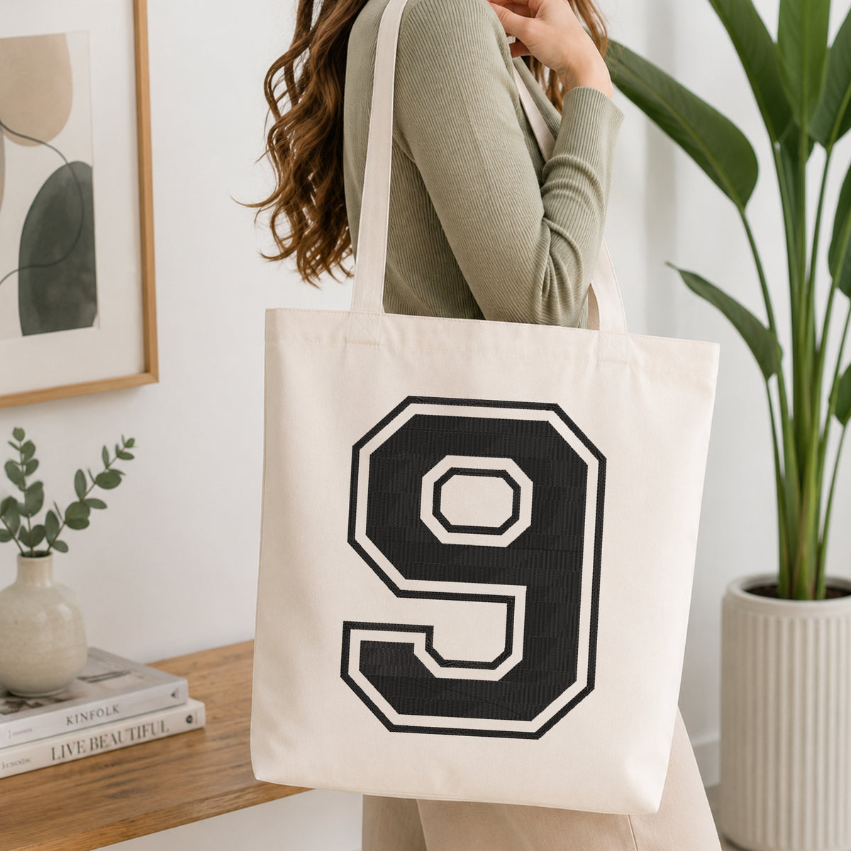 A person wearing a sage green ribbed long-sleeve top carries a cream canvas tote bag with a large black varsity-style number 9 outlined in white on the front, set in a bright, modern room with a wooden console table, framed art, and a potted plant.