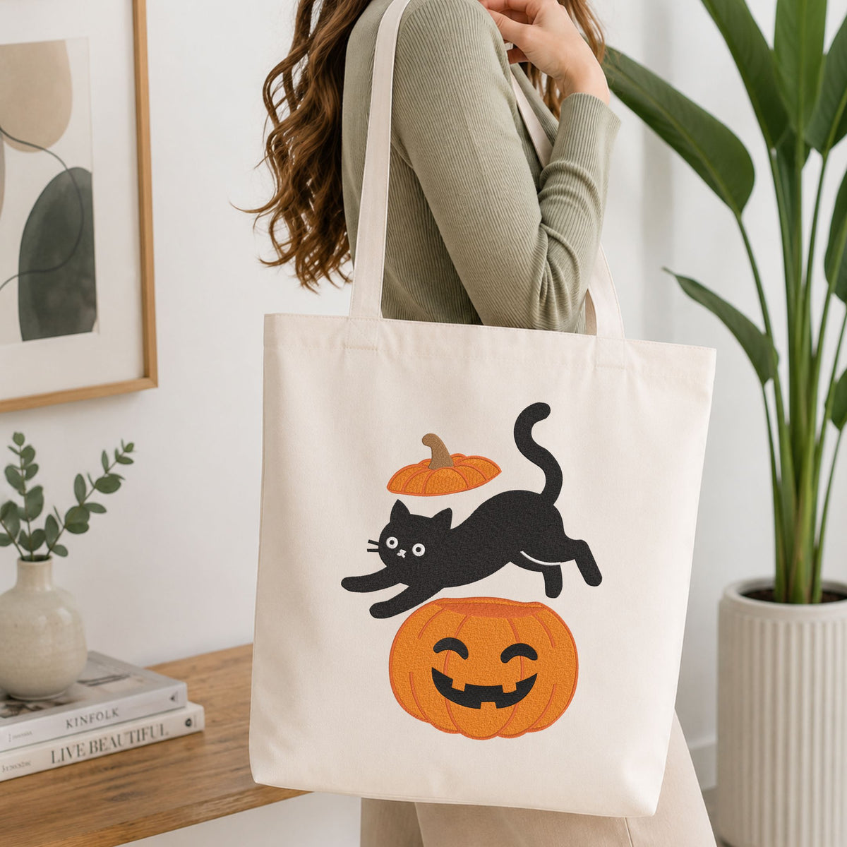 Woman carrying a white natural canvas tote bag featuring a playful black cat jumping over a jack-o-lantern pumpkin machine embroidery design in a bright, modern interior setting   