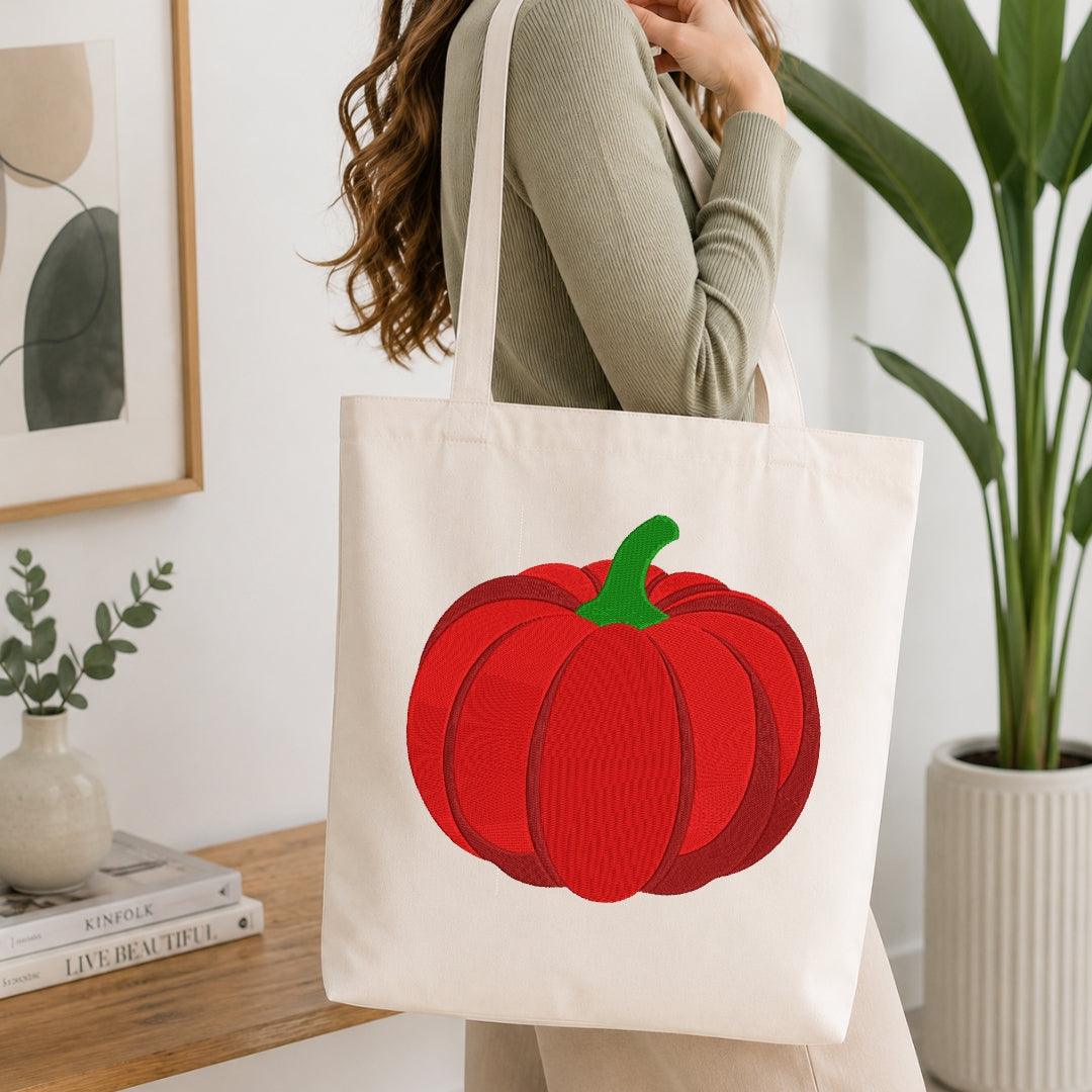 A woman wearing a sage green ribbed sweater carries a cream canvas tote bag with a large red pumpkin graphic and a green stem, set in a bright, modern room with a wooden table, books, and a potted plant.