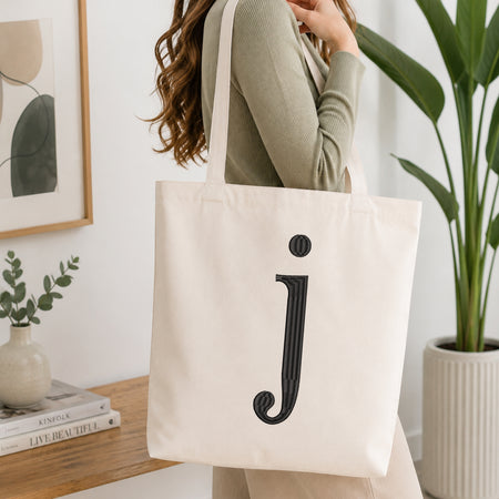 A woman wearing a sage green ribbed long-sleeve top carries a large off-white canvas tote bag with a black embroidered lowercase 'j' on the front, set in a bright, modern room with a wooden table, framed art, and a potted plant.