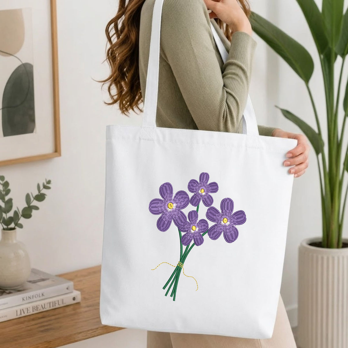 Woman carrying a white natural canvas tote bag featuring an embroidered purple flowers bouquet design in a bright, modern interior setting