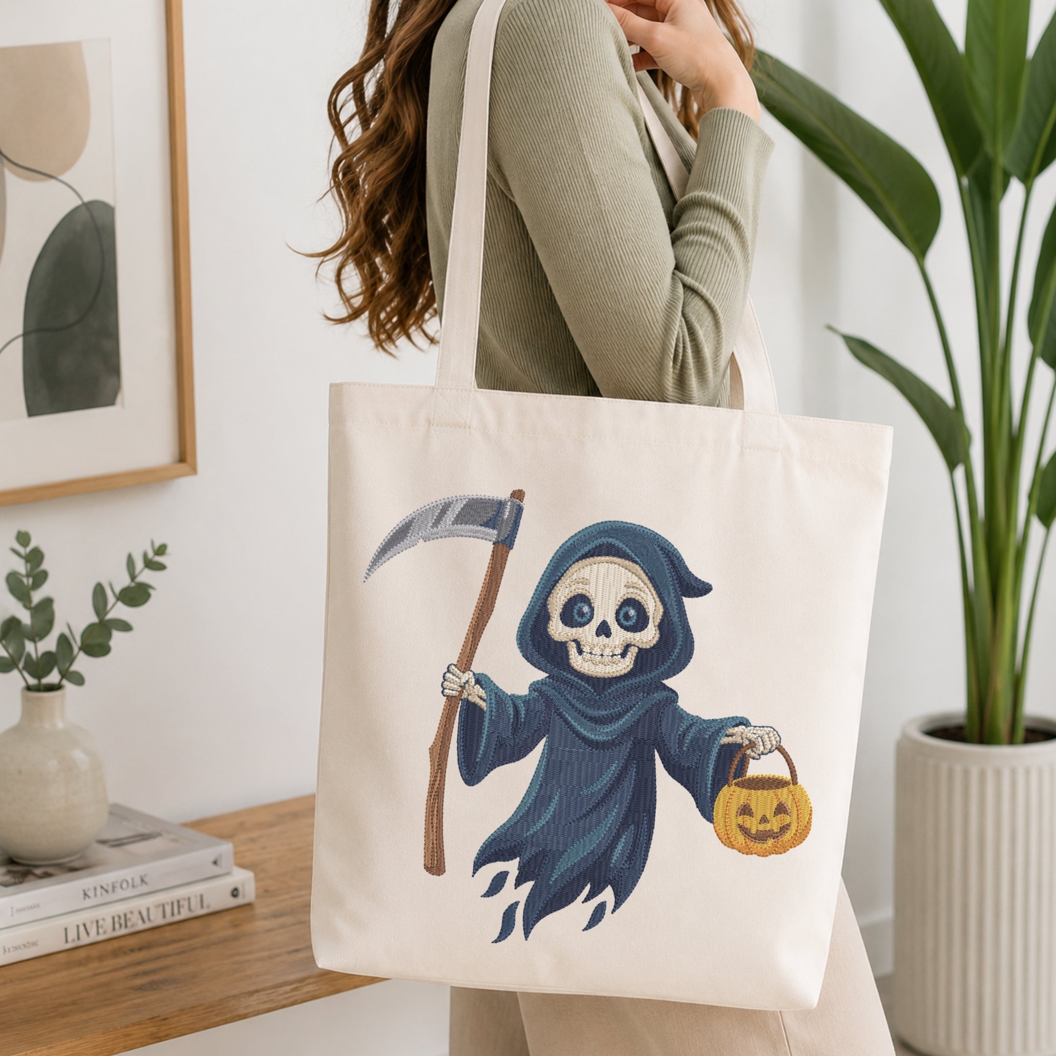 A woman wearing a sage green top carries a cream canvas tote bag featuring a cartoon grim reaper illustration holding a scythe and a jack-o'-lantern.
