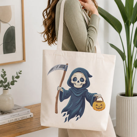 A woman wearing a sage green top carries a cream canvas tote bag featuring a cartoon grim reaper illustration holding a scythe and a jack-o'-lantern.