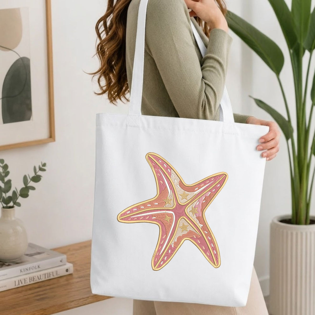Folded white cotton t-shirt mockup featuring a pink and beige coastal starfish machine embroidery design, styled with cotton stems and a wooden cup.