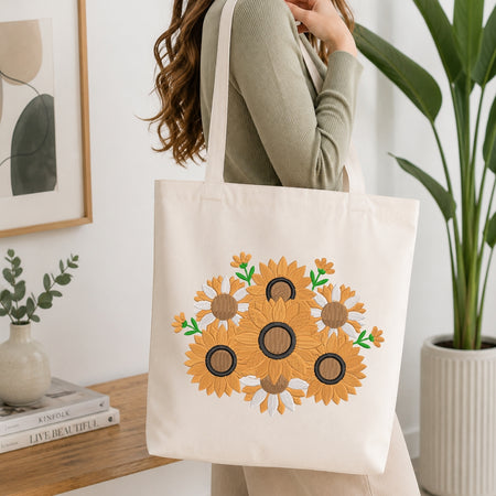 A person wearing a sage green ribbed sweater carries a cream canvas tote bag decorated with a large embroidered sunflower design in yellow, brown, and green. The scene is set in a bright, modern room with a wooden table, books, a framed print, and a potted plant.