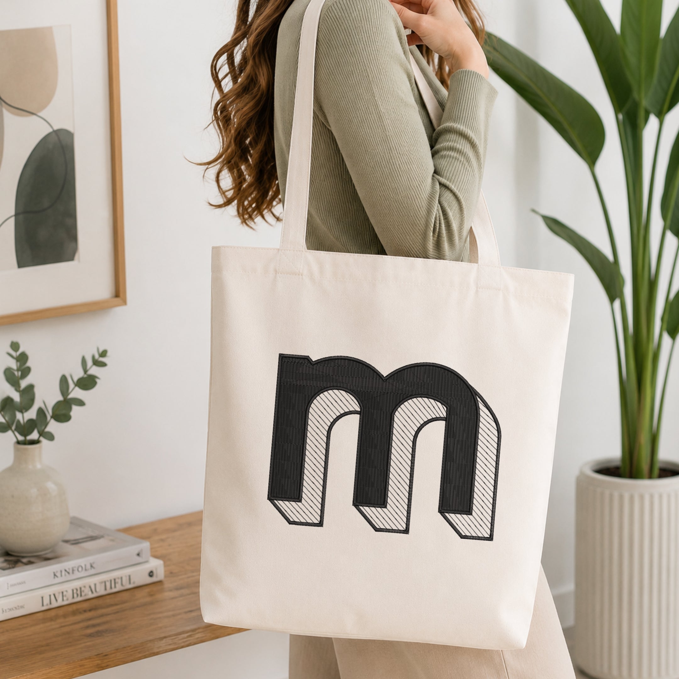 A woman wearing a sage green ribbed long-sleeve top carries a cream canvas tote bag with a large black embroidered letter 'm' on the front, set in a bright, modern room with a wooden console table, framed art, and a potted plant.