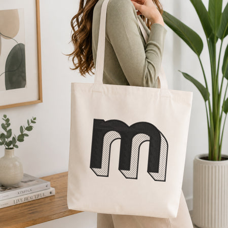 A woman wearing a sage green ribbed long-sleeve top carries a cream canvas tote bag with a large black embroidered letter 'm' on the front, set in a bright, modern room with a wooden console table, framed art, and a potted plant.