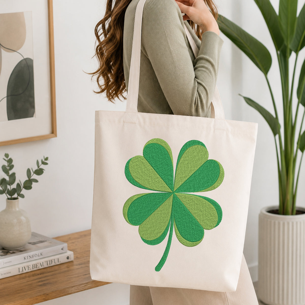 A woman wearing a sage green ribbed sweater carries a cream canvas tote bag with a large green four-leaf clover design on the front, set in a bright, modern room with a wooden table, framed art, and a potted plant.