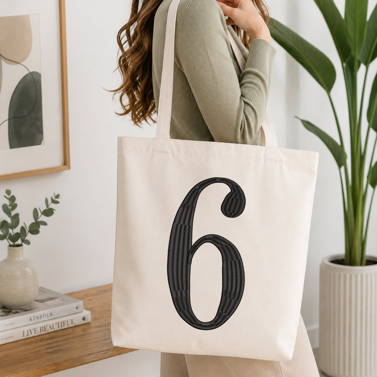 A woman wearing a sage green ribbed top carries a large off-white canvas tote bag with a bold black striped number 6 printed on the front, set in a bright, modern room with a wooden console table and a potted plant.
