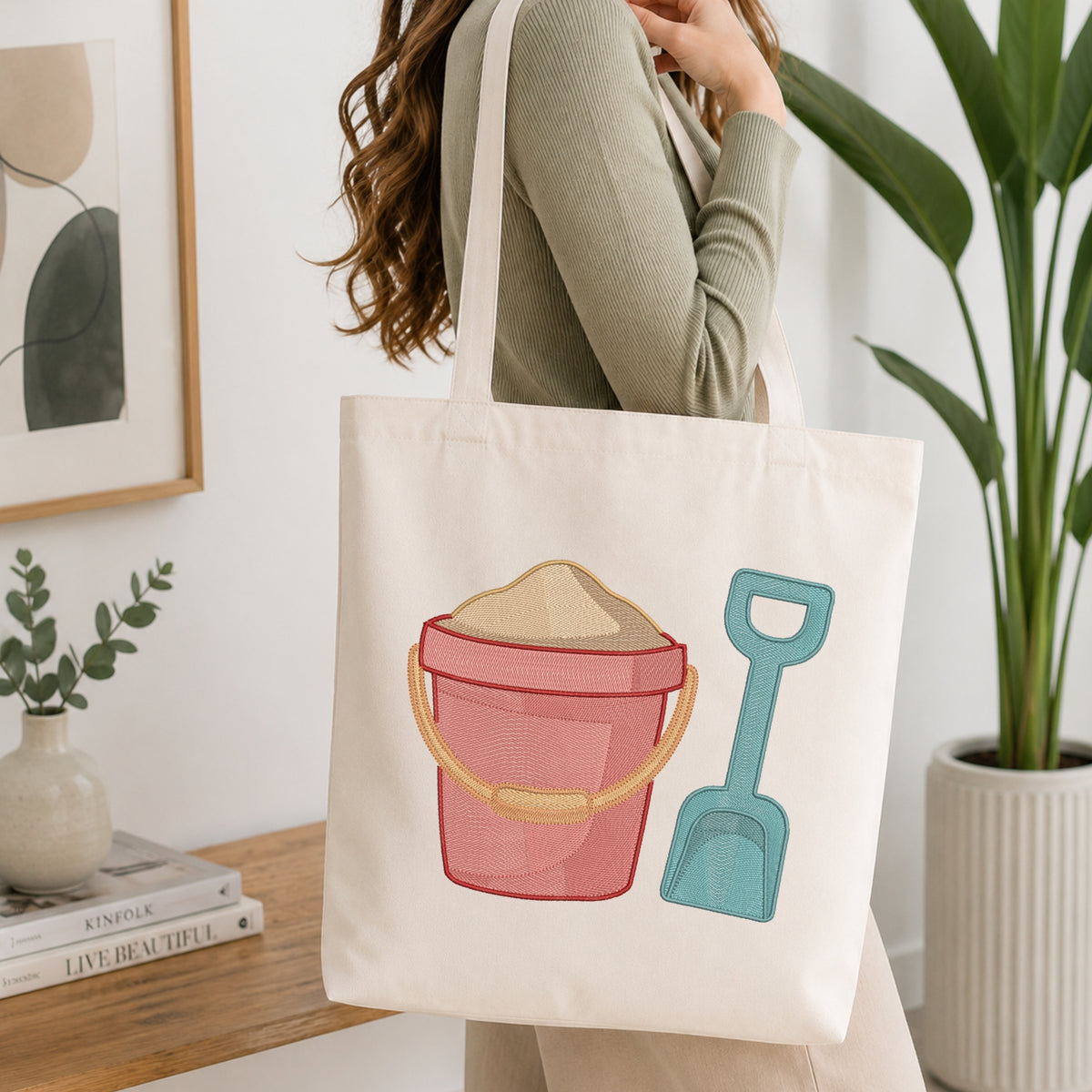 A woman holds a cream canvas tote bag featuring a colorful embroidered design of a pink bucket with sand and a teal shovel, set against a bright, modern indoor background with a potted plant and framed art.