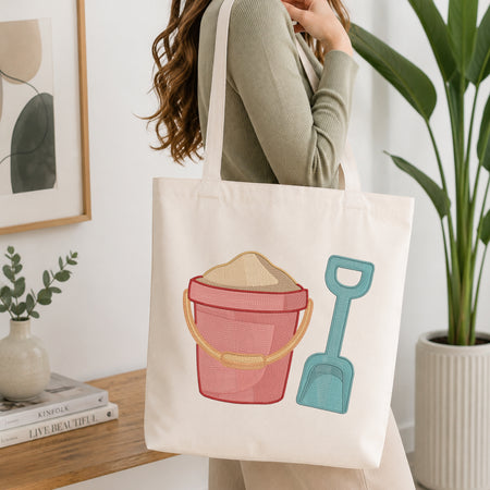 A woman holds a cream canvas tote bag featuring a colorful embroidered design of a pink bucket with sand and a teal shovel, set against a bright, modern indoor background with a potted plant and framed art.