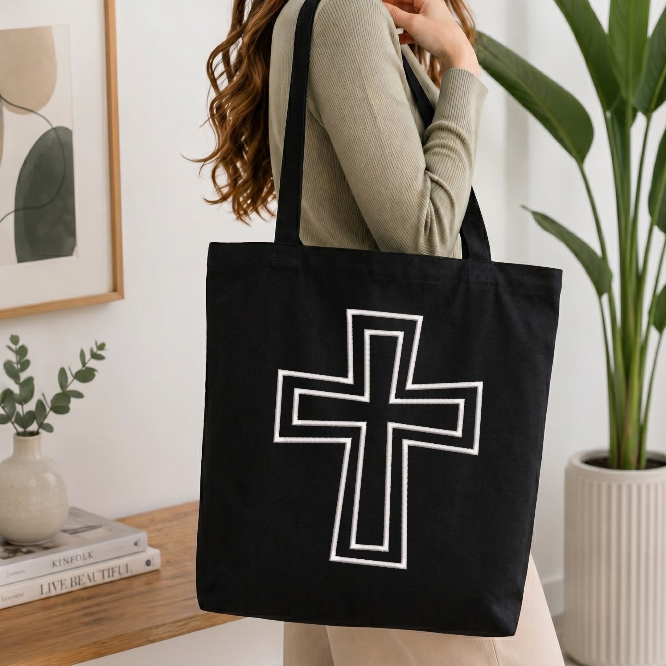 A person wearing a green top holds up a black canvas tote bag printed with the white outlined Christian cross embroidery design, set against a bright, minimalist room background with indoor plants.