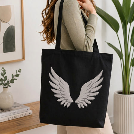 A woman wearing a beige ribbed sweater carries a black canvas tote bag with a large white embroidered angel wings design on the front, set against a bright, modern indoor background with a potted plant and framed art.
