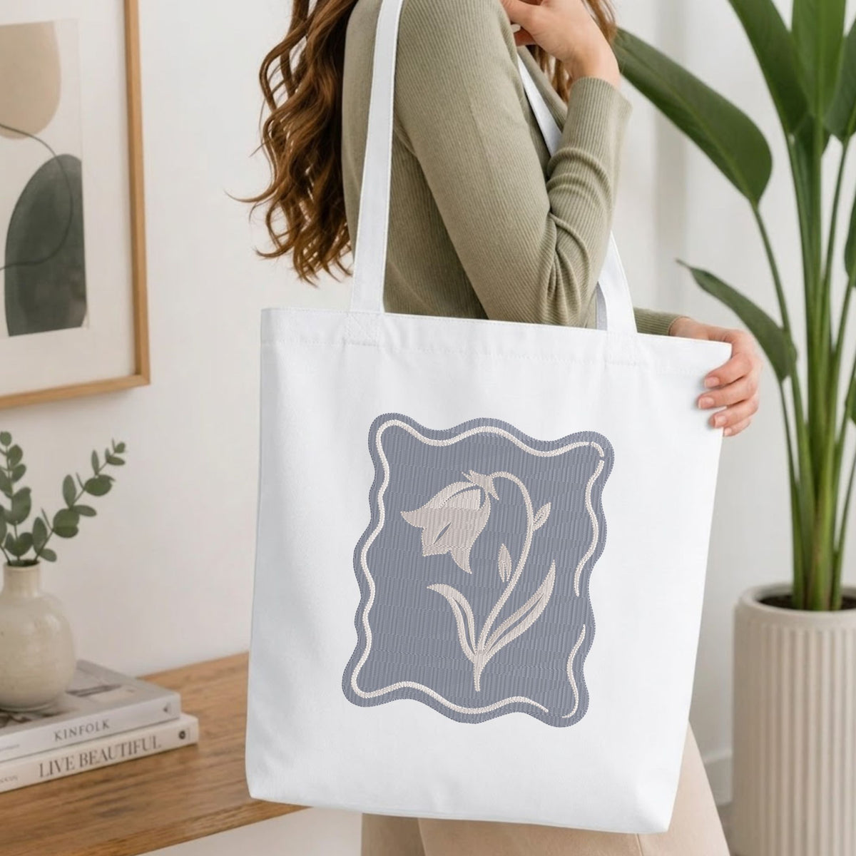 Woman carrying a white natural canvas tote bag featuring an embroidered purple bellflower design in a bright, modern interior setting.   