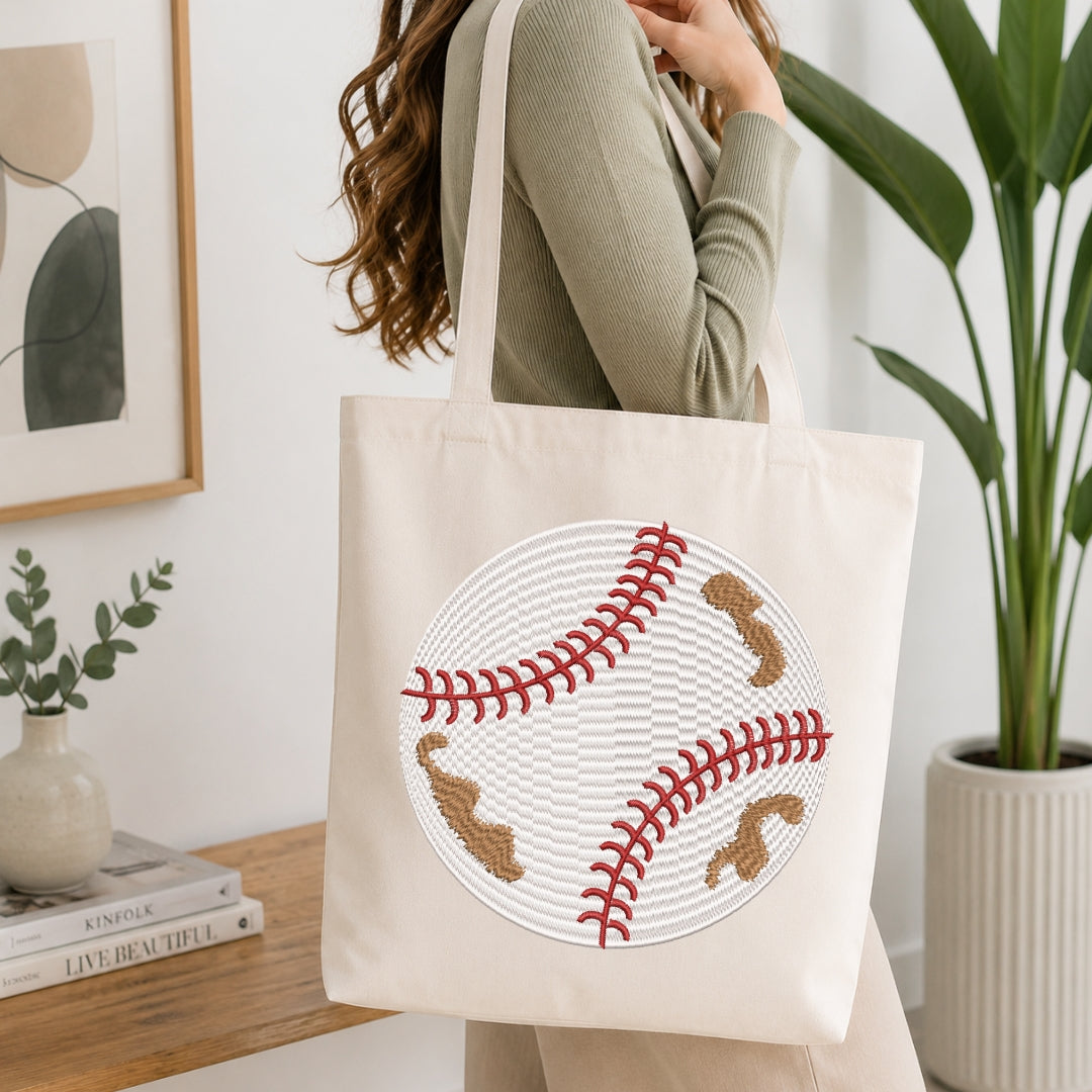 A woman carries a cream canvas tote bag featuring a large embroidered baseball design with red stitching and brown patches, set against a bright, modern indoor background with a wooden table and potted plants.