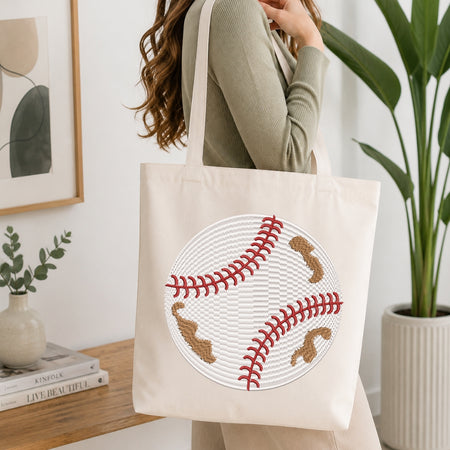 A woman carries a cream canvas tote bag featuring a large embroidered baseball design with red stitching and brown patches, set against a bright, modern indoor background with a wooden table and potted plants.