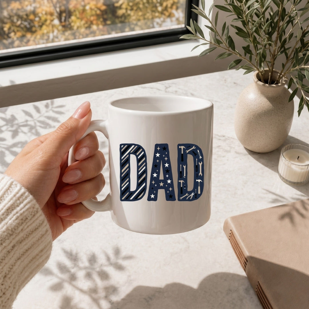 Hand holding a white mug with 'DAD' printed on it, placed on a table with a window and plant in the background.
