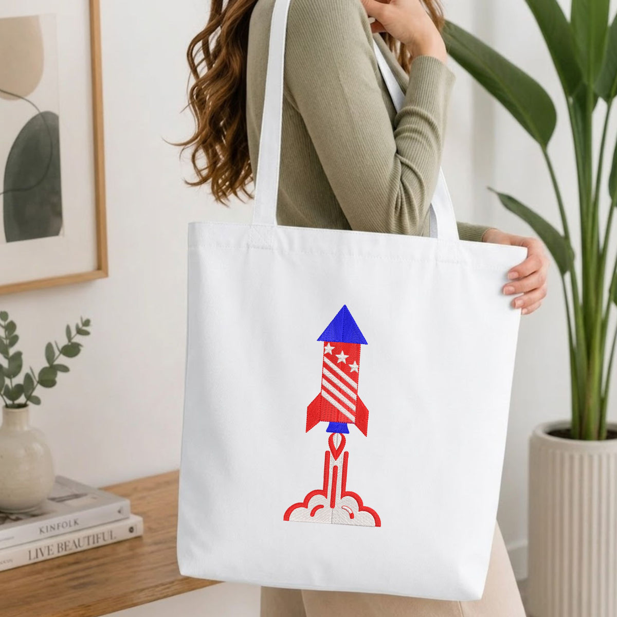 Woman carrying a white natural canvas tote bag featuring an embroidered patriotic space rocket design in a bright, modern interior setting.