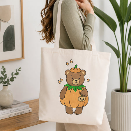 A person wearing a sage green ribbed long-sleeve top carries a cream canvas tote bag. The bag features a cute cartoon illustration of a brown teddy bear dressed in a pumpkin costume with candy corn accents.