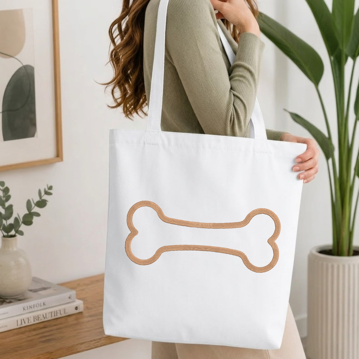 A woman wearing a sage green ribbed sweater carries a white canvas tote bag with a large tan bone outline graphic on the front, set in a bright, modern room with a wooden table and potted plants.