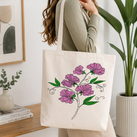 An off-white canvas tote bag with a purple and green floral print, carried by a woman in a ribbed sage green top in a bright, modern room.