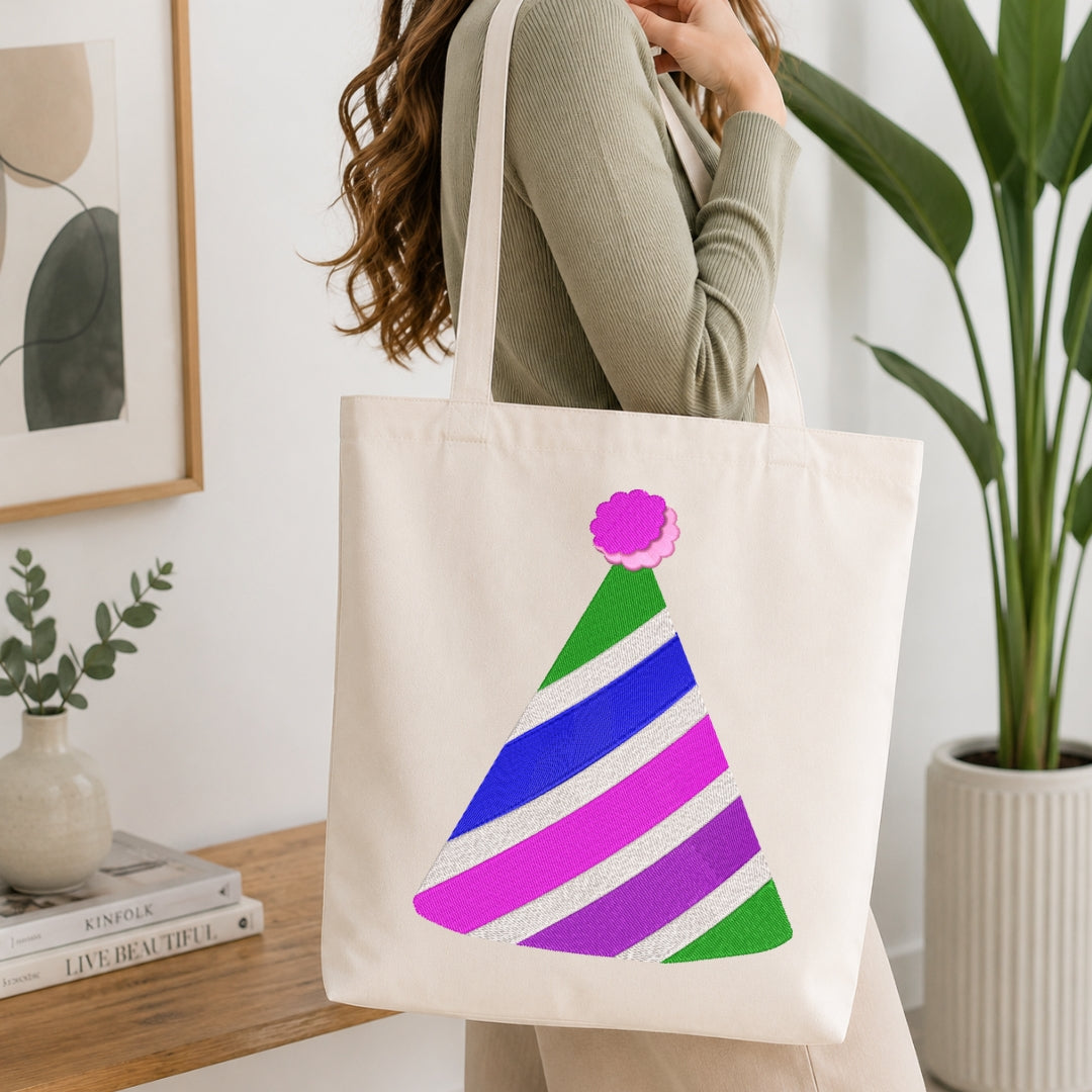 A woman wearing a sage green ribbed sweater carries a cream canvas tote bag with a colorful striped party hat graphic. The background shows a cozy indoor setting with a wooden table, books, a framed print, and a potted plant.