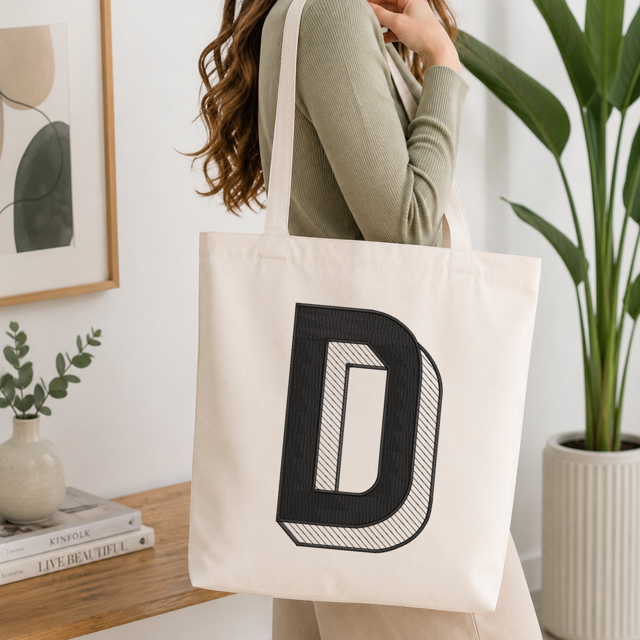 A woman wearing a sage green ribbed long-sleeve top carries a cream canvas tote bag with a large black block letter 'D' printed on the front, set in a bright, modern room with a wooden table, framed art, and a potted plant.