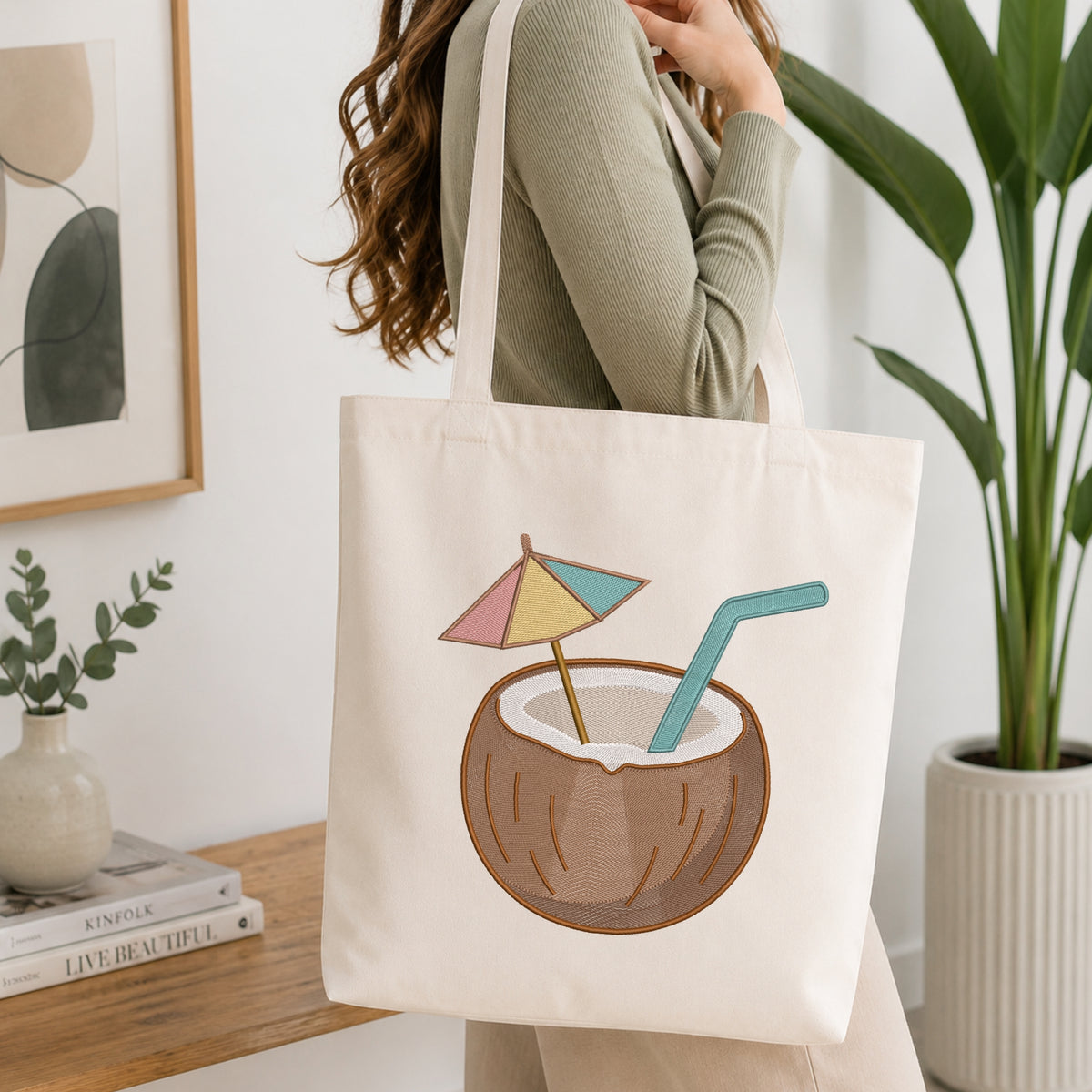 A woman wearing a sage green ribbed long-sleeve top carries a cream canvas tote bag. The bag features a graphic illustration of a coconut drink with a pastel pink, yellow, and blue beach umbrella and a teal straw.