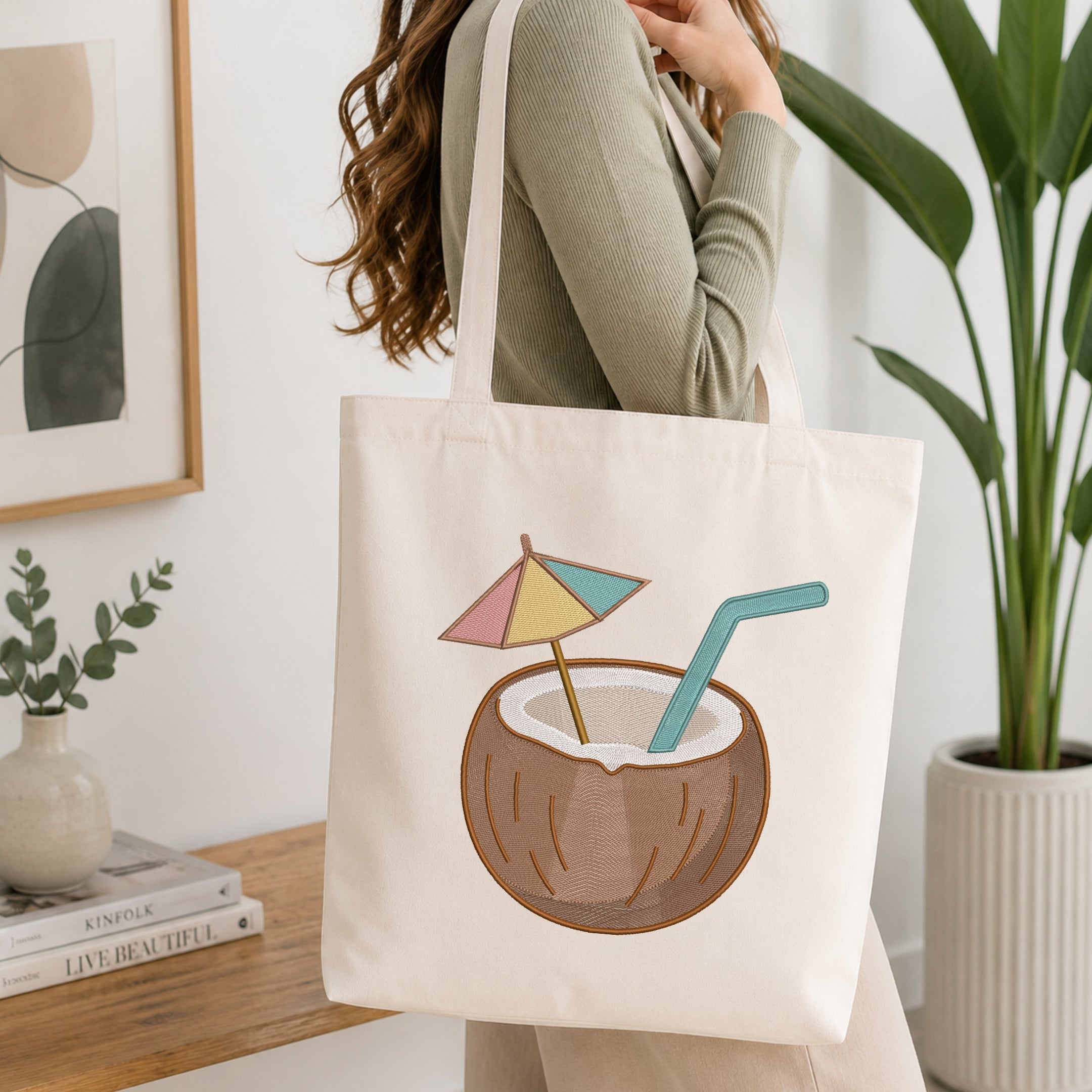 A woman wearing a sage green ribbed long-sleeve top carries a cream canvas tote bag. The bag features a graphic illustration of a coconut drink with a pastel pink, yellow, and blue beach umbrella and a teal straw.