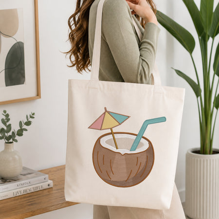 A woman wearing a sage green ribbed long-sleeve top carries a cream canvas tote bag. The bag features a graphic illustration of a coconut drink with a pastel pink, yellow, and blue beach umbrella and a teal straw.