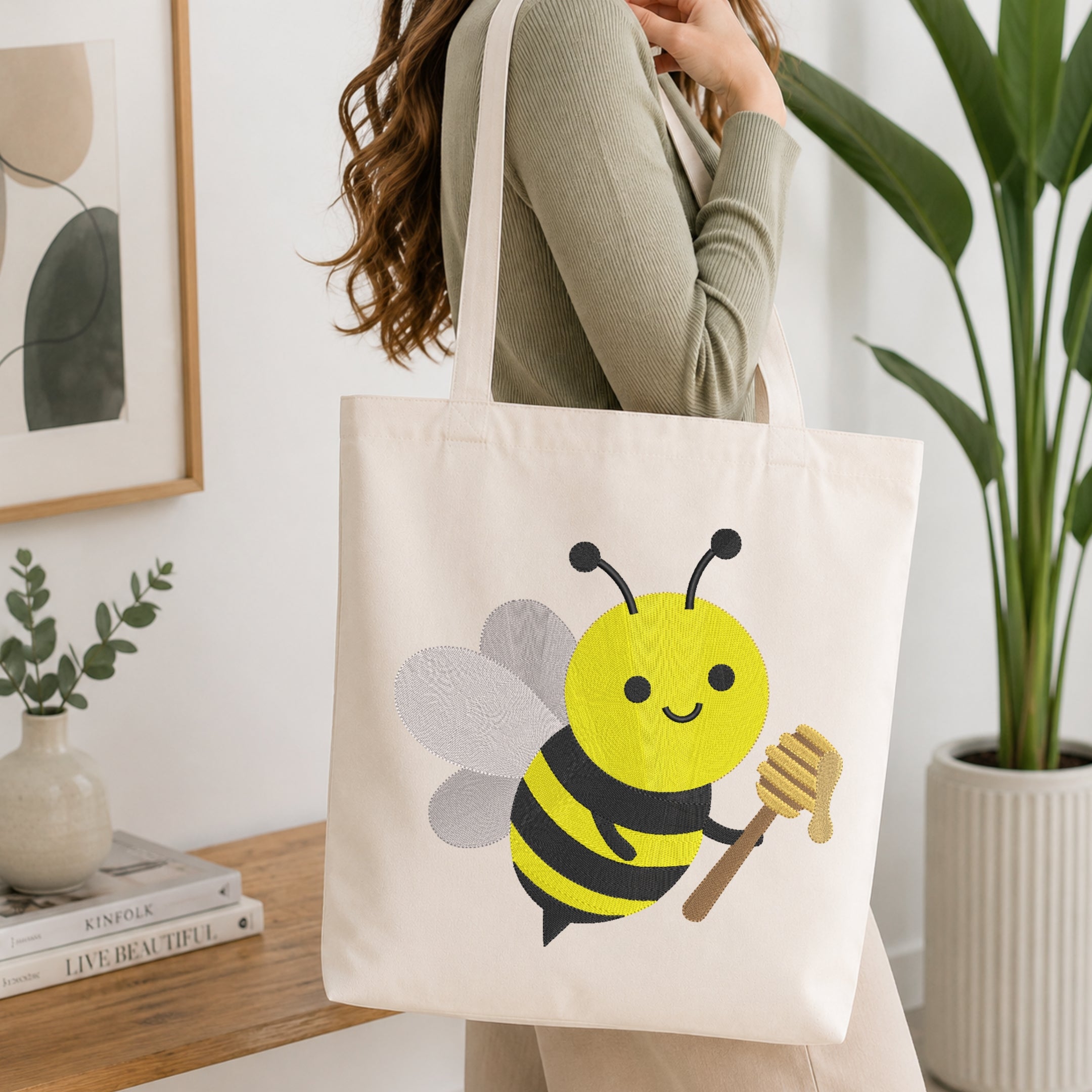 A woman wearing a sage green ribbed sweater carries a cream canvas tote bag with a cute yellow and black embroidered bee holding a honeycomb on the front. The scene is set in a bright, modern room with a wooden shelf, books, a framed print, and a potted green plant.
