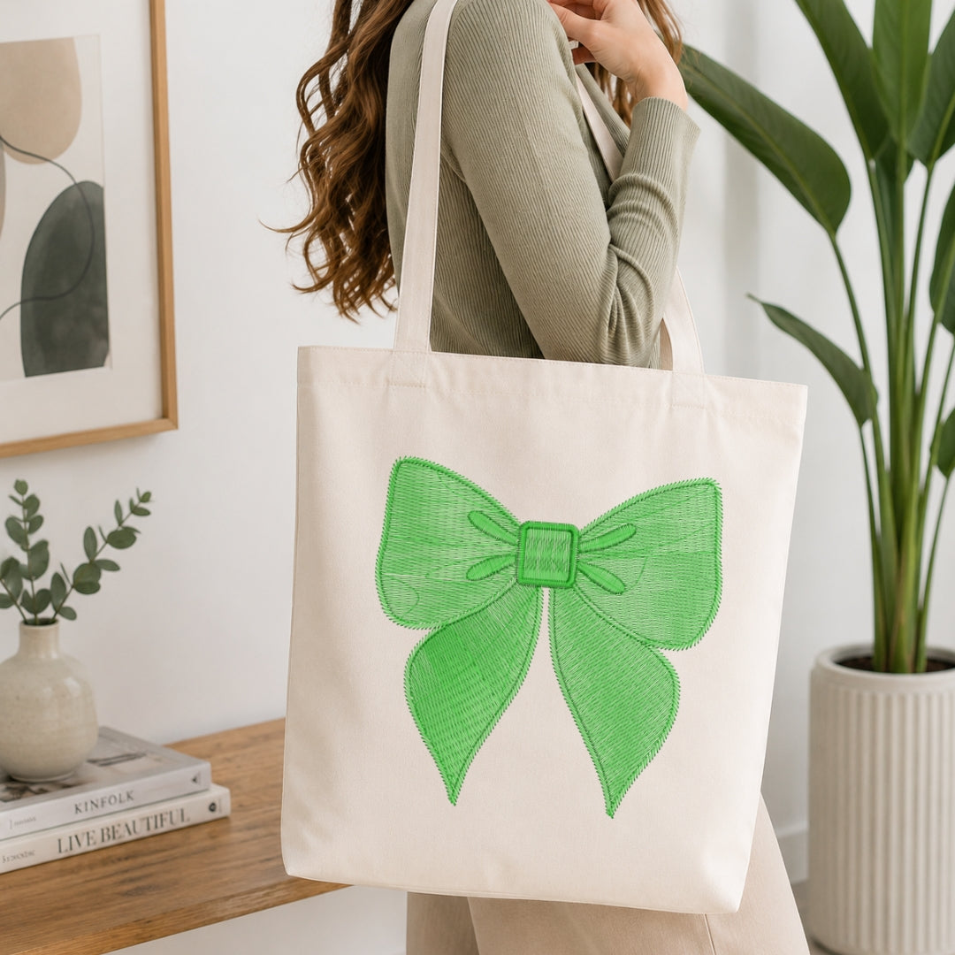 A person wearing a ribbed sage green top carries a cream canvas tote bag with a large green bow graphic on the front, set in a bright, modern room with a wooden table, books, and a potted plant.