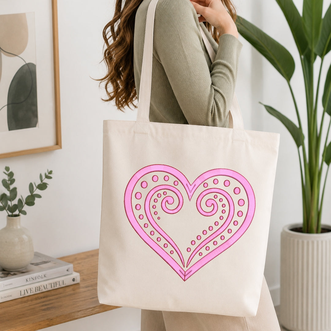 A woman wearing a sage green ribbed sweater carries a cream canvas tote bag with a large pink and magenta heart design printed on the front, set in a bright, modern room with a wooden table, books, and a potted plant.