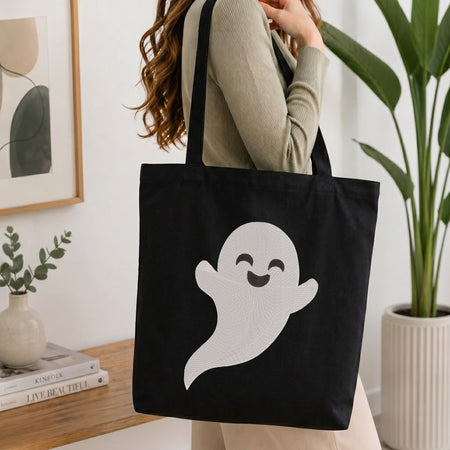 Woman carrying a black natural canvas tote bag featuring an embroidered cute smiling ghost design in a bright, modern interior setting.  