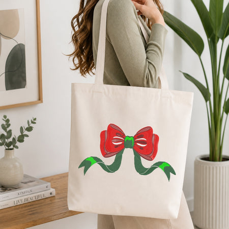 A woman wearing a ribbed sage green long-sleeve top carries a cream canvas tote bag with a red bow and green ribbon graphic printed on the front, set in a bright, modern room with indoor plants.