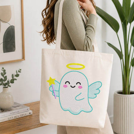 A person wearing a sage green ribbed sweater carries a cream canvas tote bag with a cute embroidered angel design. The bag features a light blue outlined angel with a yellow halo, pink cheeks, and a purple wand.