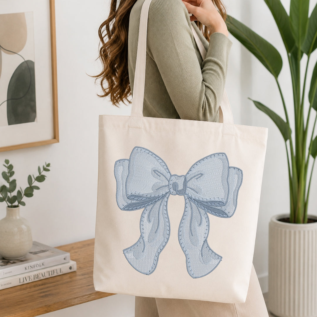 An off-white canvas tote bag with a large light blue bow graphic, carried by a person wearing a sage green ribbed sweater in a bright, modern room with a wooden table and potted plants.