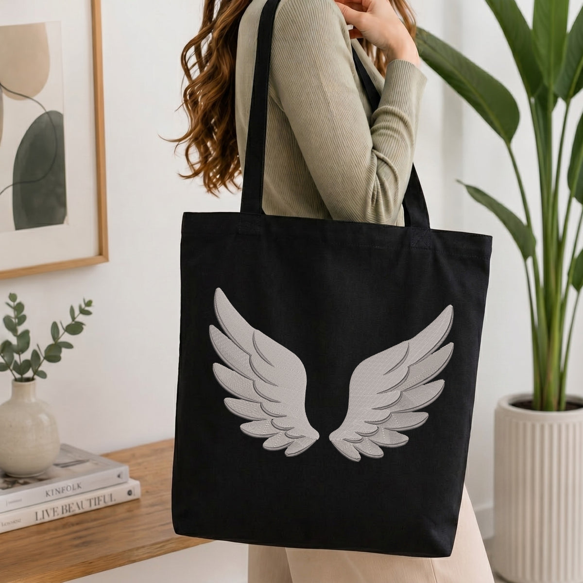 A woman wearing a beige ribbed sweater carries a black canvas tote bag with a large white embroidered angel wings design on the front, set in a bright, modern room with a wooden table, framed art, and a potted plant.