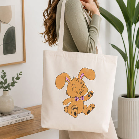 Woman carrying a white natural canvas tote bag featuring an embroidered cute golden-brown bunny design in a bright, modern interior setting.