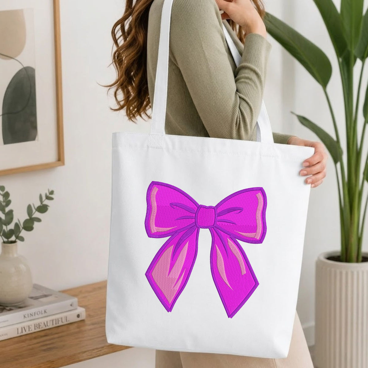Woman carrying a white natural canvas tote bag featuring an embroidered vibrant purple bow design in a bright, modern interior setting.